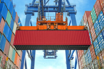 Red container lifted by an orange and blue crane in a container ship yard