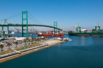 Cargo ships docked at a busy port with a large green suspension bridge and container cranes under a clear blue sky.