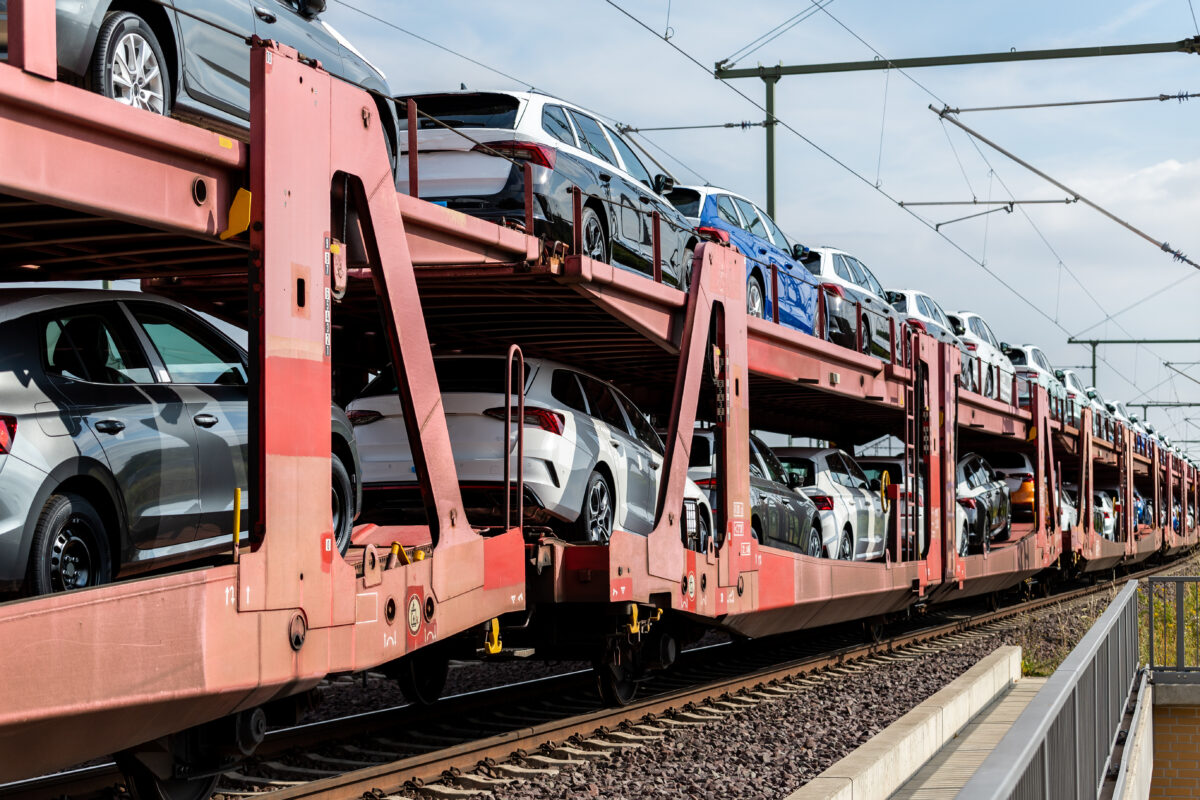 Multi-level rail freight cars loaded with new cars are parked on railway tracks under overhead electric lines, showing rows of vehicles secured on red transport wagons.