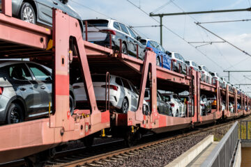 Multi-level rail freight cars loaded with new cars are parked on railway tracks under overhead electric lines, showing rows of vehicles secured on red transport wagons.